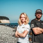 Eine Frau in einem weißen Poloshirts neben einem Mann im schwarzen T-Shirt mit einem Cap auf dem Kopf vor dem Kreidefelsen auf der Ostseeinsel Rügen.