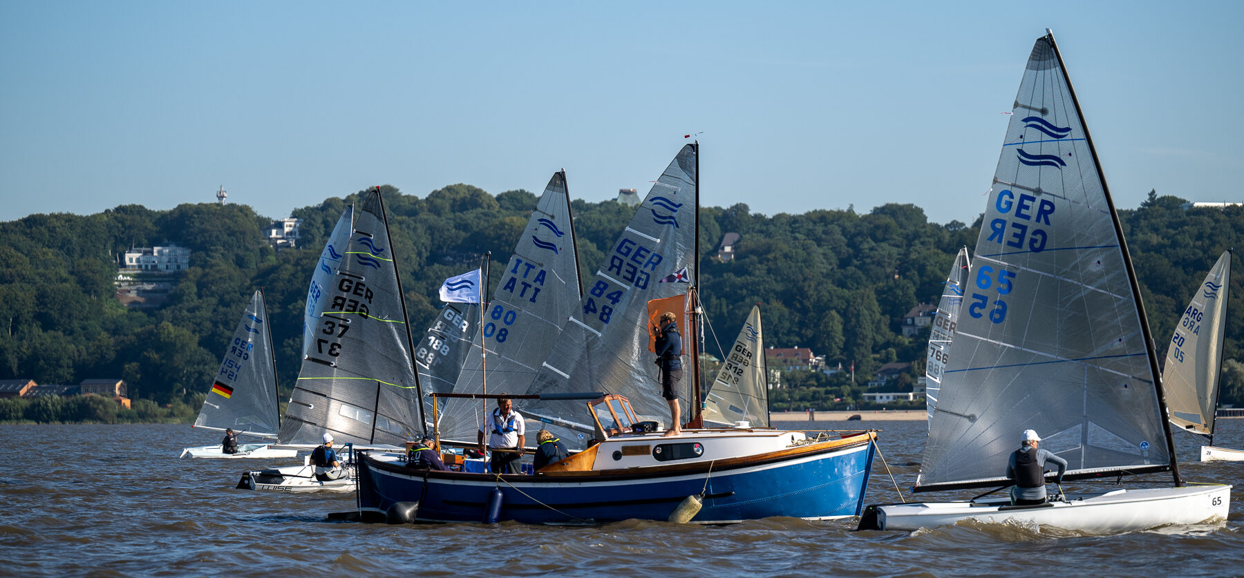 Jollen vom Typ Finn-Dinghy starten auf der Elbe in eine Regatta. Im Hintergrund ist der Hamburger Stadtteil Blankenese zu sehen