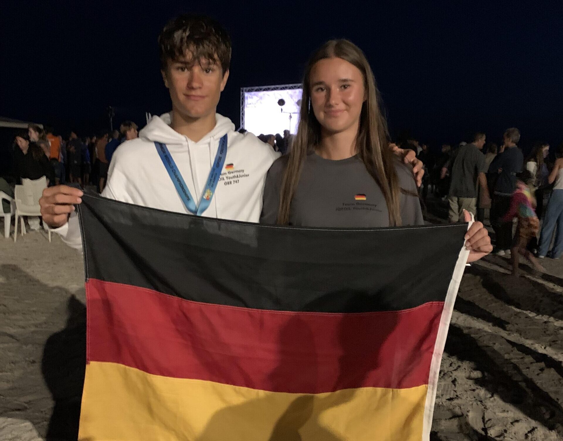 Die Geschwister Moritz und Emma Schleicher halten lächelnd eine Deutschland-Fahne hoch, der Hintergrund ist dunkel