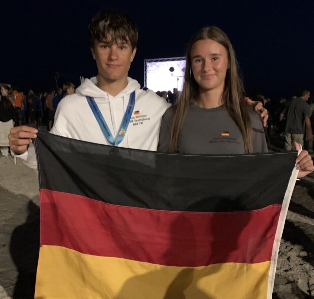 Die Geschwister Moritz und Emma Schleicher halten lächelnd eine Deutschland-Fahne hoch, der Hintergrund ist dunkel