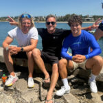 Jannis Maus, DSV-Trainer Aleksei Chibizov und Jan Vöster sitzen auf einer Mauer. Sie lächeln in die Kamera. Im Hintergrund ist das Meer zu sehen