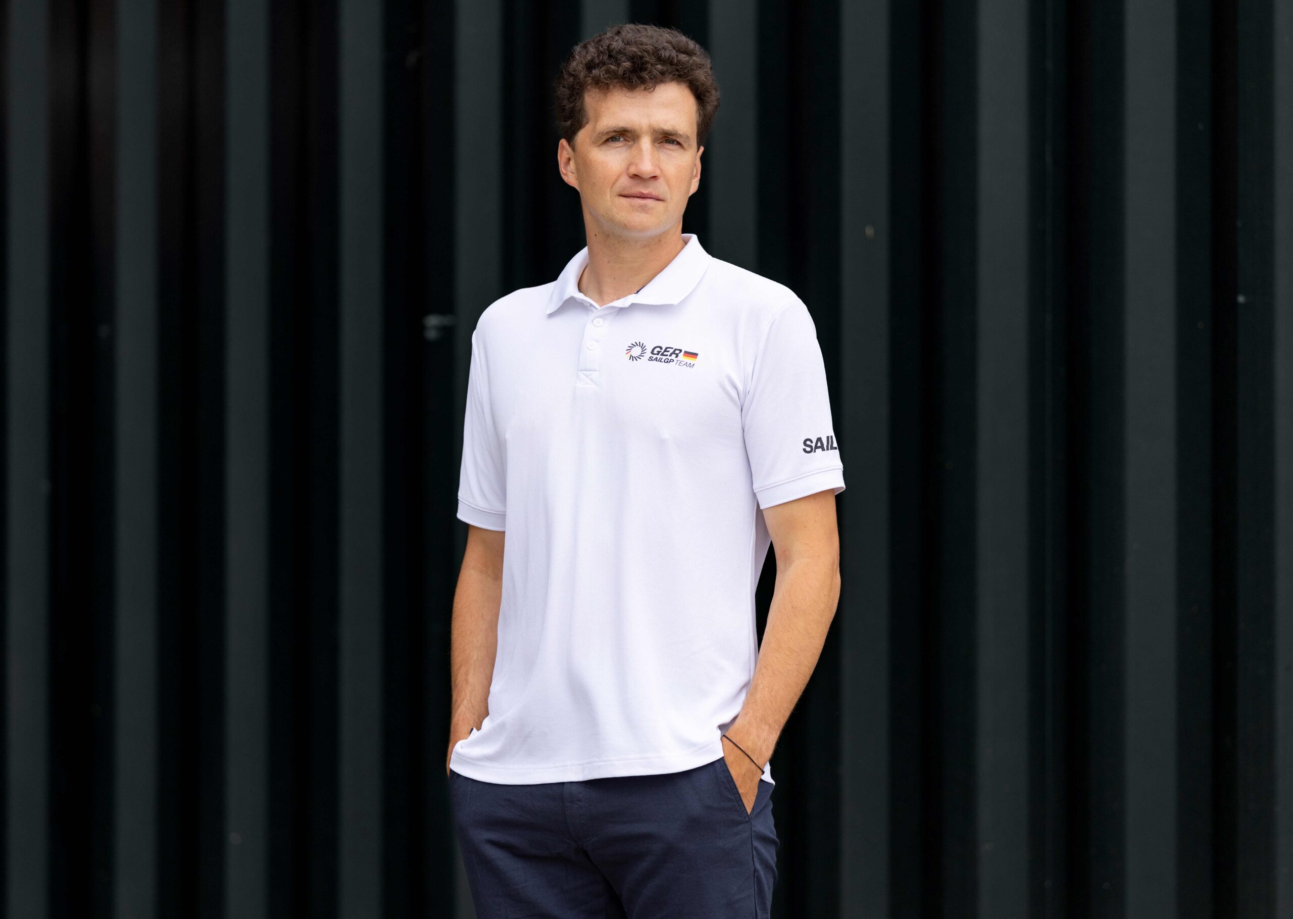 Ein dunkelhaariger Mann mit weißem Poloshirt guckt in die Kamera