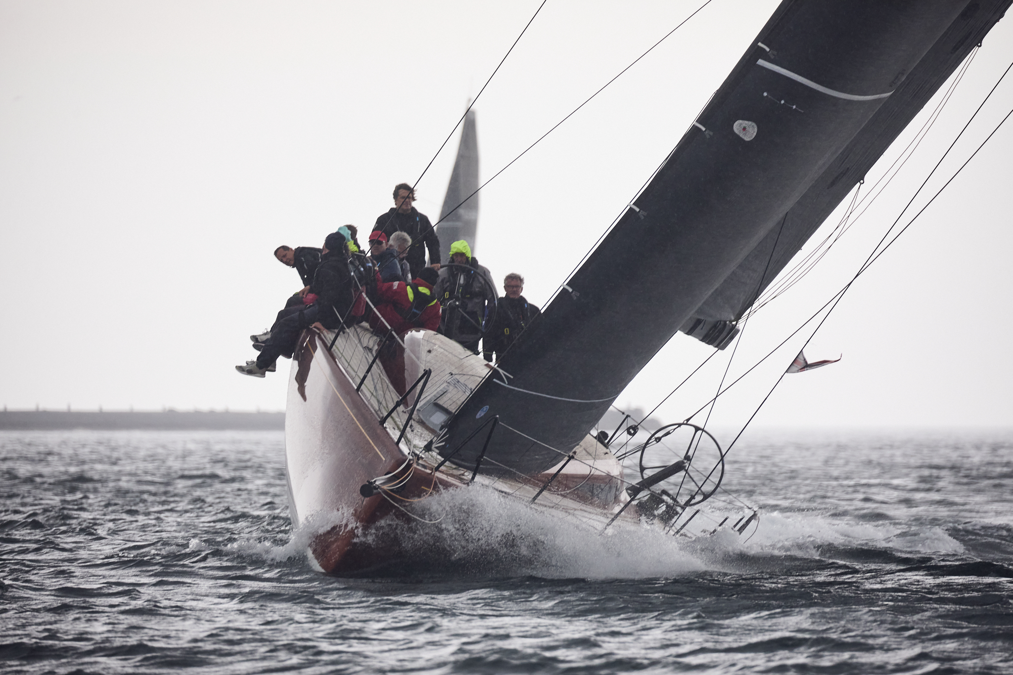 Eine Yacht mit hölzernem Rumpf segelt bei viel Wind auf Kreuzkrus, die Crew sitzt auf der Kante auf der Steuerbordseite.