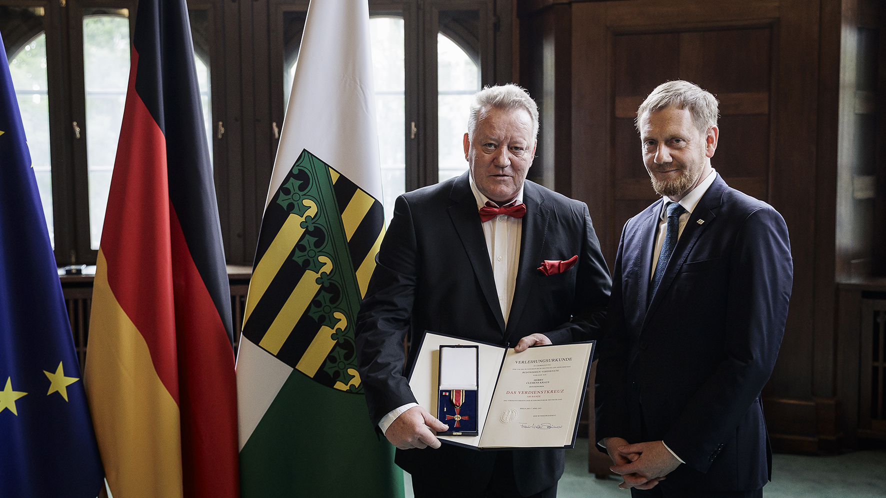 Clemens Kraus steht neben dem Ministerpräsidenten von Sachsen, Michael Kretschmer, in der Staatskanzlei in Dresden. Im Hintergrund sind die Flaggen von Sachsen, Deutschland und der EU zu sehen. Clemens Kraus trägt am Revers das Bundesverdienstkreuz und hält in den Händen eine Urkunde.