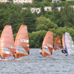 Viele Windsurfer flitzen über einen See, die meisten Segel sind orange, nur eines ist transparent. Es wehr ein starker Wind.