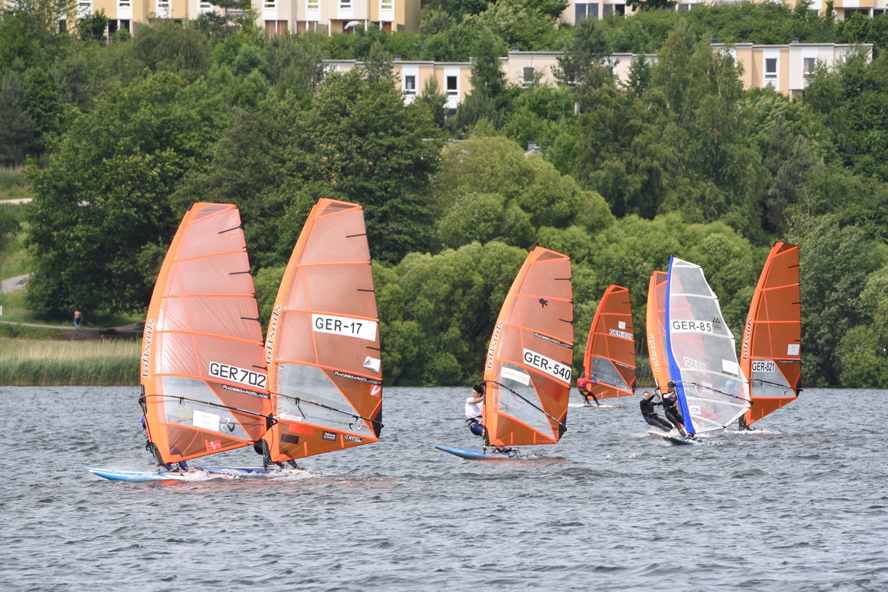Viele Windsurfer flitzen über einen See, die meisten Segel sind orange, nur eines ist transparent. Es wehr ein starker Wind.