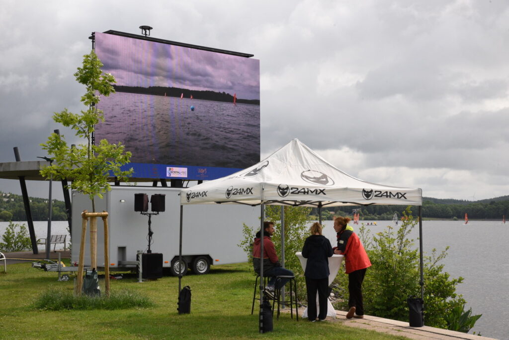 Auf einer Leinwand kann man die Surfwettfahrten auf dem Wasser beobachten. Die große Wand steht an Land am einem See, dem Bostalsee.
