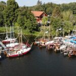 Vor einem Grundstück mit viel Wald ragen zwei Stege in einen See. An den Stegen liegen viele Segelboote. Der Himmel ist blau, die Sonne scheint.
