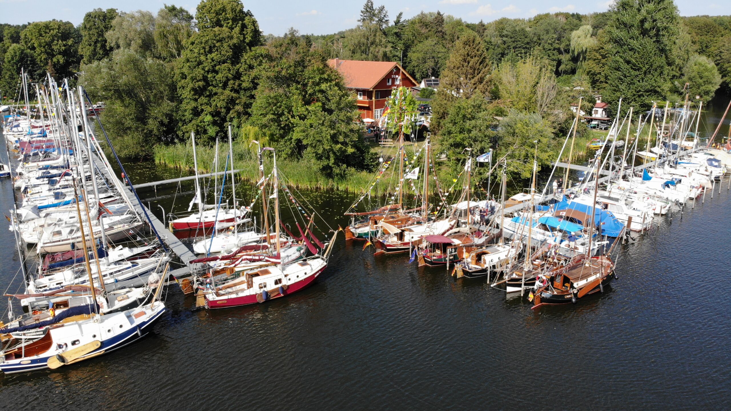 Vor einem Grundstück mit viel Wald ragen zwei Stege in einen See. An den Stegen liegen viele Segelboote. Der Himmel ist blau, die Sonne scheint.
