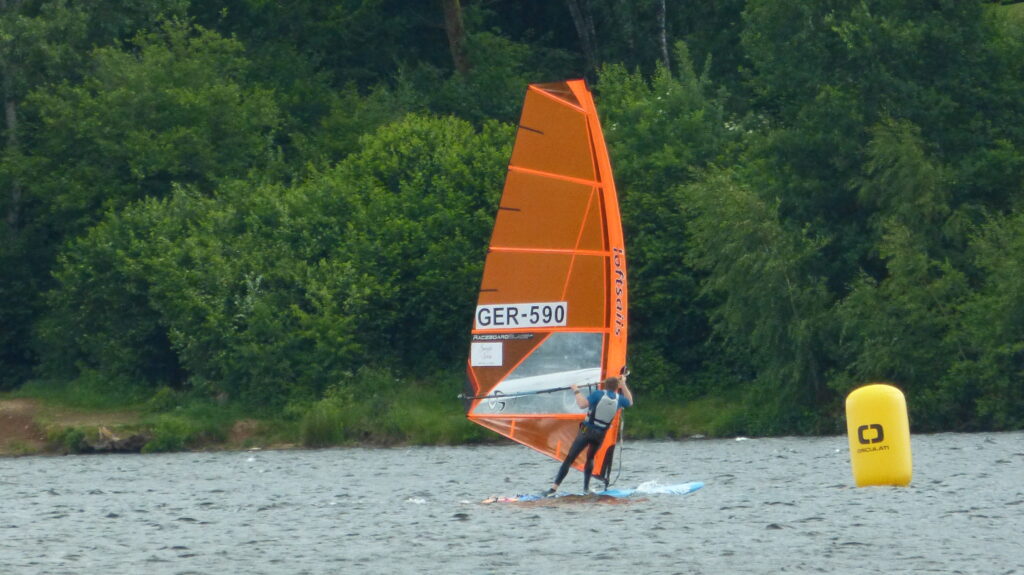 Der Deutsche Meister Fabian Aldinger surft auf einem langen Windsurfbrett, das Segel ist orange und hat die Segelnummer GER 590. Rechts liegt eine gelbe Tonne