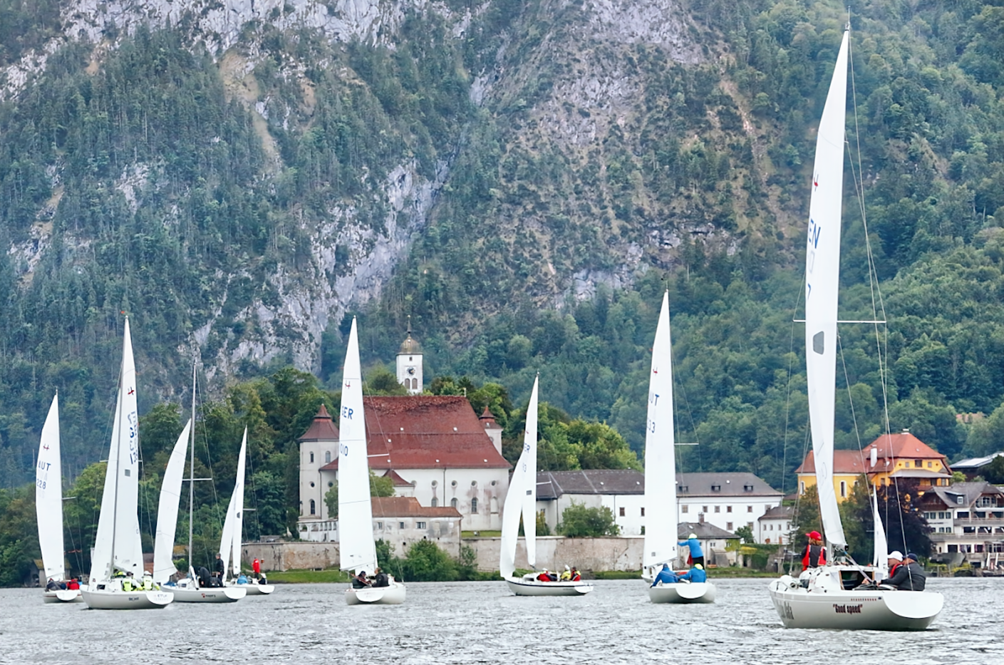 Hohe Felswand und davor der Traunsee mit segelnden H-Booten auf denen Seglerinnen und Segler zu erkennen sind.