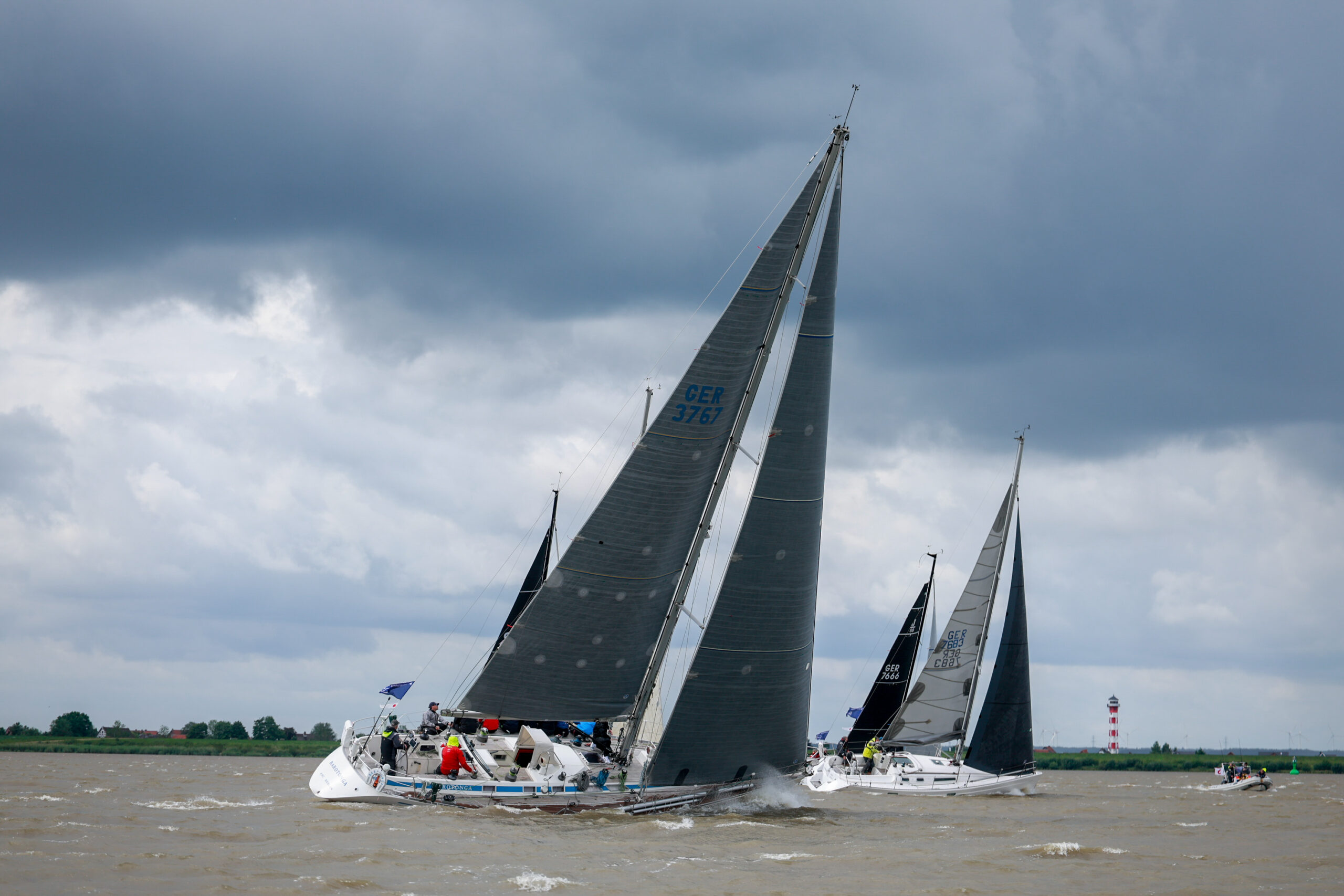 Eine Yacht auf der Elbe bei einer Regatta mit viel Wind, Regen und Welle. Im Hintergrund ein rot-weißer Leuchtturm zwei weitere Yachten und ein Schlauchboot.