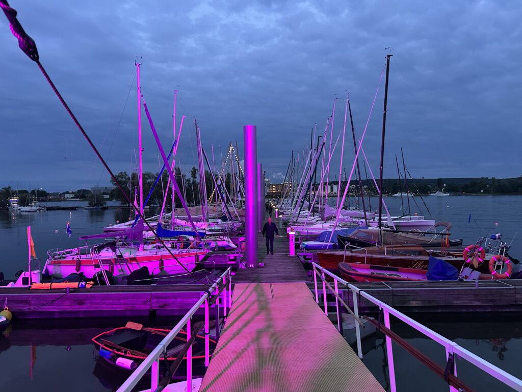 Ein Bootssteg in abendlichem Licht, Scheinwerfer leuchten ihn zusätzlich lila an. Viele Boote sind am Steg festgemacht, einige sind geschmückt.