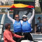 Marla Bergmann und Hanna Wille stehen im bauen Bib auf ihrem Boot und halten jubelnd eine Deutschlandflagge in die Luft. Vor ihnen sitzt ihr Coach Maurice Paardenkooper auf dem Motorboot