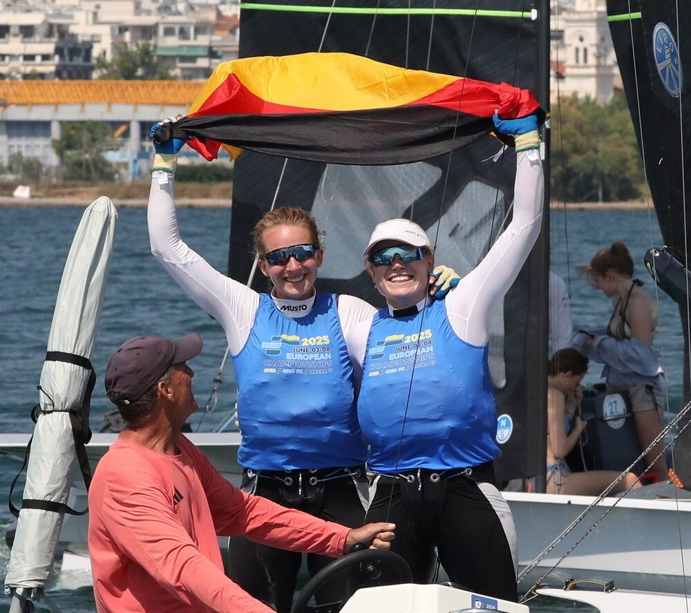 Marla Bergmann und Hanna Wille stehen im bauen Bib auf ihrem Boot und halten jubelnd eine Deutschlandflagge in die Luft. Vor ihnen sitzt ihr Coach Maurice Paardenkooper auf dem Motorboot