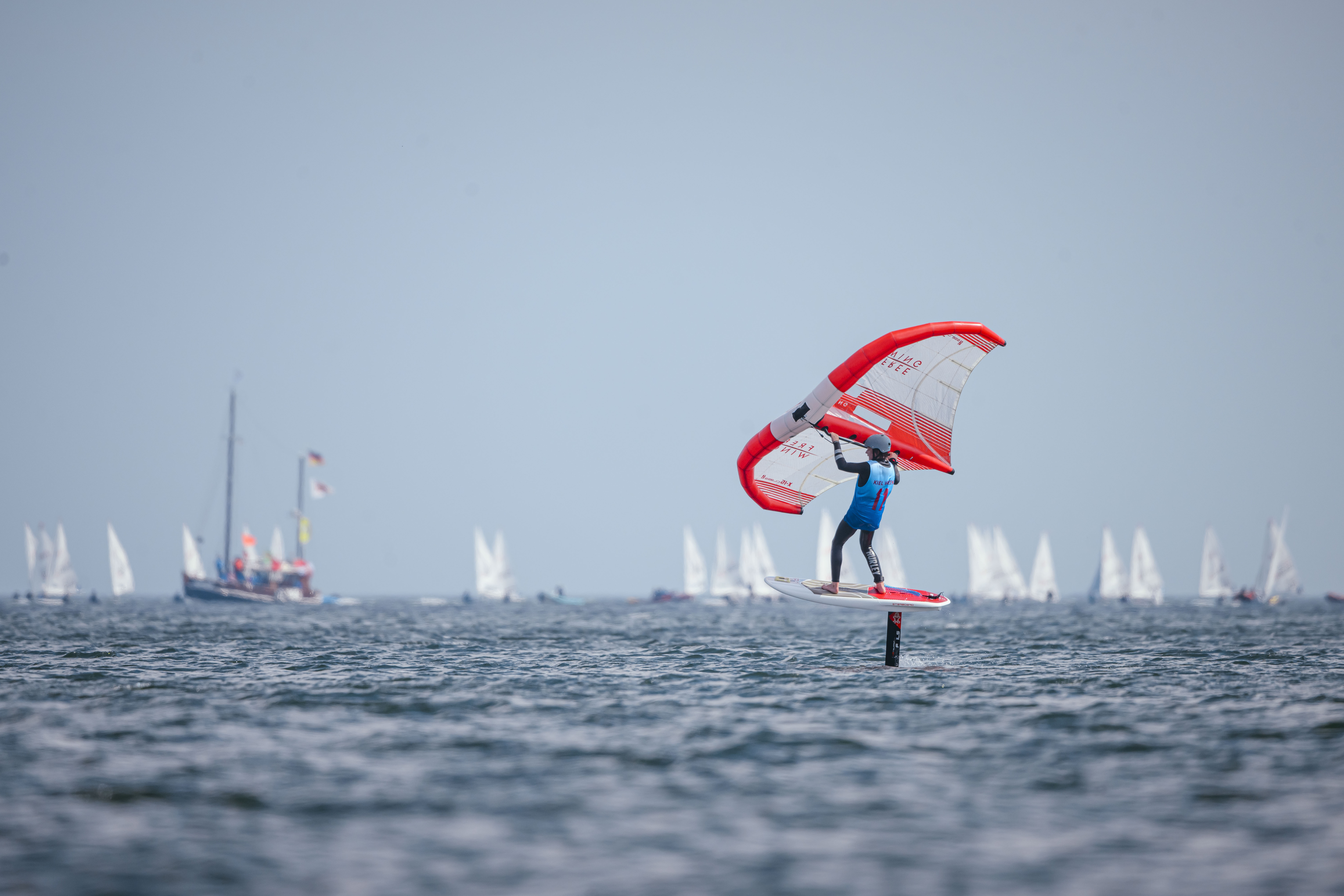 Ein Wingfoiler schwebt über die Kieler Förde. Im Hintergrund sind Segelschiffe zu sehen.