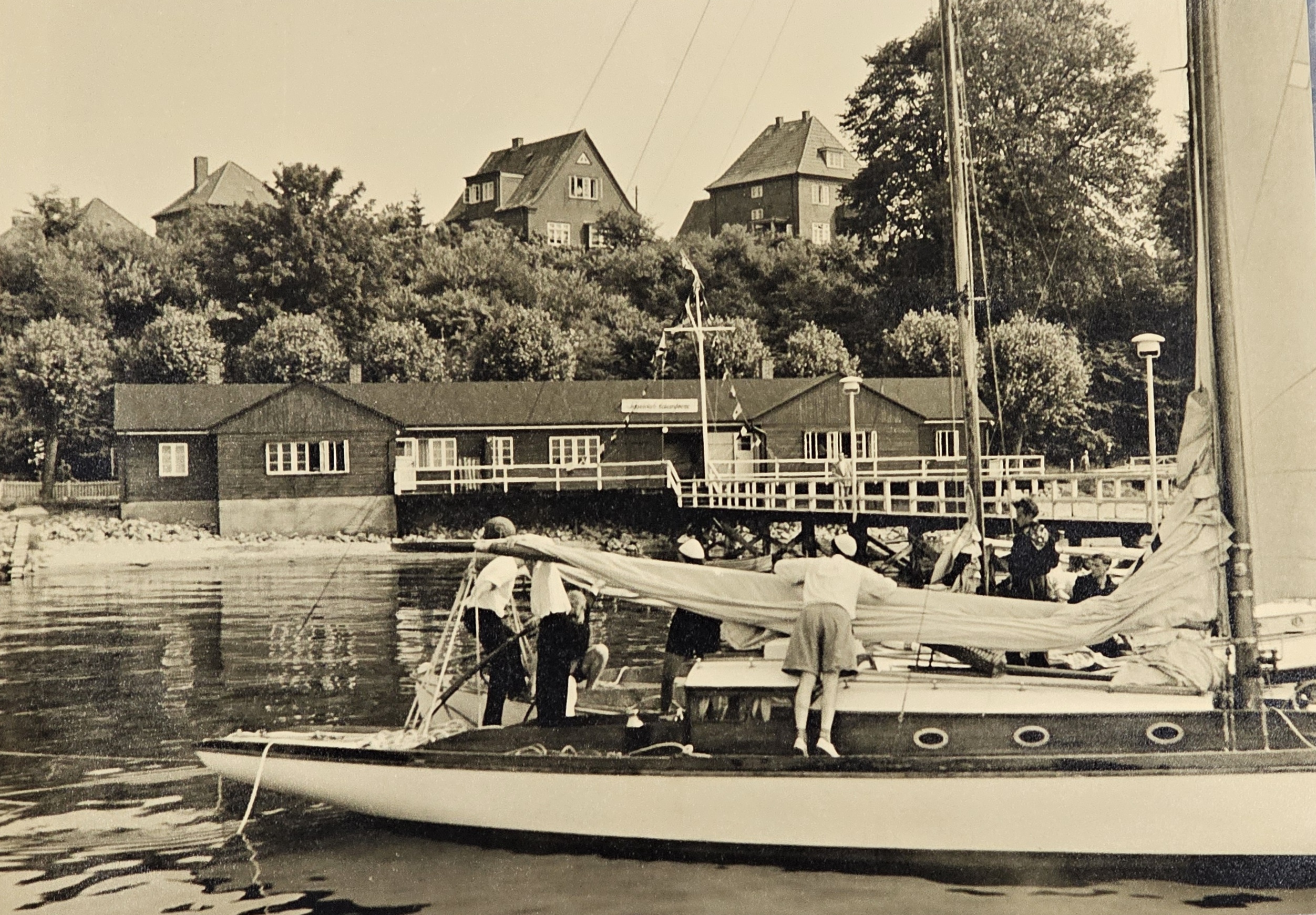 Ein altes Clubhaus eines Segelvereins mit einer Yacht davor auf der Menschen ein Großsegel auftuchen.