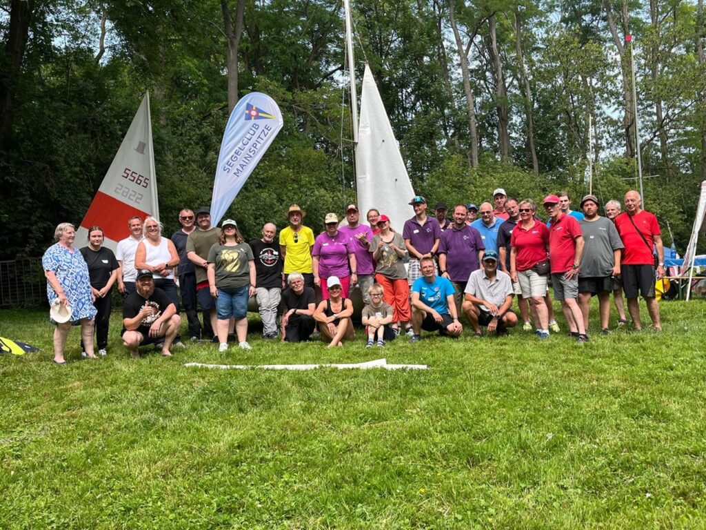 Das Foto zeigt eine Gruppe von Kindern, Jugendlichen und Erwachsenen beim Segelclub Mainspitze. Sie stehen und sitzen auf dem Rasen und wollen gemeinsam das Segeln lernen.