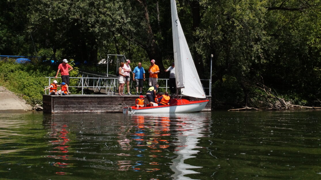 Menschen stehen auf einem Steg. Davor liegt ein Boot im Wasser mit zwei hochgezogenen Segeln. Im Boot sitzen Kinder mit Schwimmwesten, ein Mann zeigt ihnen, wie an Bord alles bedient wird