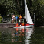 Menschen stehen auf einem Steg. Davor liegt ein Boot im Wasser mit zwei hochgezogenen Segeln. Im Boot sitzen Kinder mit Schwimmwesten, ein Mann zeigt ihnen, wie an Bord alles bedient wird
