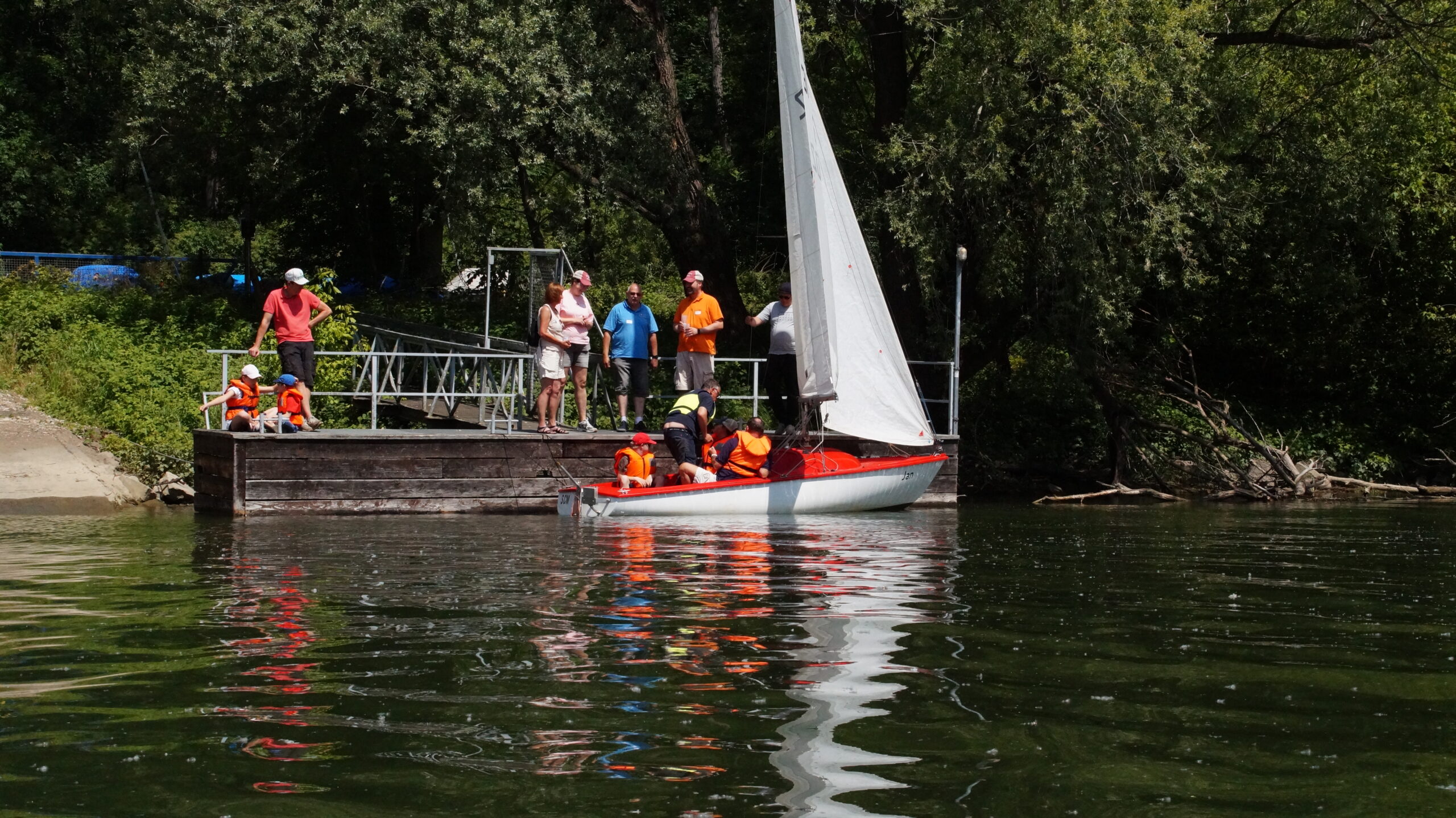 Menschen stehen auf einem Steg. Davor liegt ein Boot im Wasser mit zwei hochgezogenen Segeln. Im Boot sitzen Kinder mit Schwimmwesten, ein Mann zeigt ihnen, wie an Bord alles bedient wird