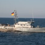 Ein graues Minenjagdboot der Deutschen Marine mit deutscher Flagge auf einem Signalmast in der Mitte des Schiffes.
