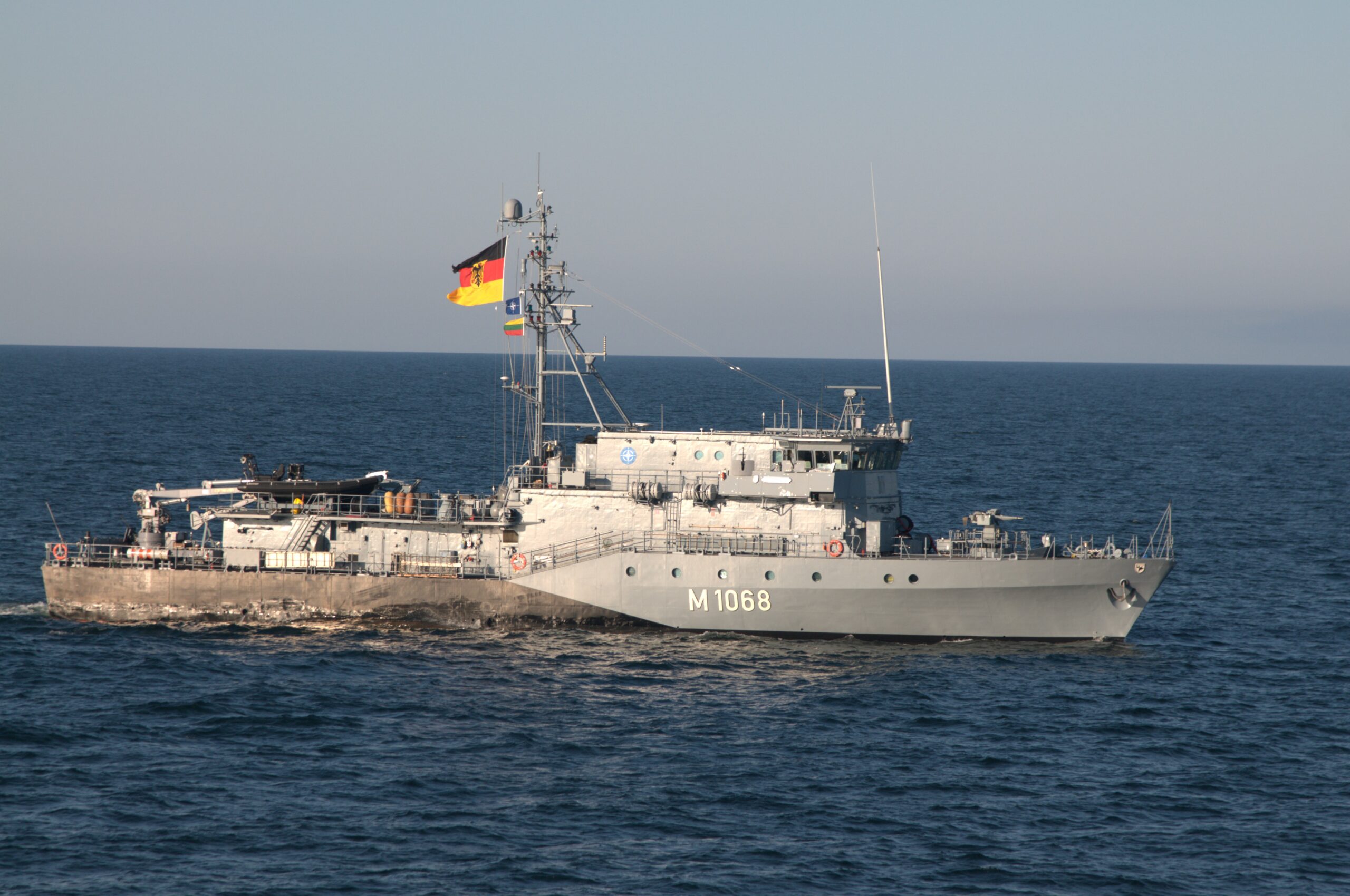 Ein graues Minenjagdboot der Deutschen Marine mit deutscher Flagge auf einem Signalmast in der Mitte des Schiffes.