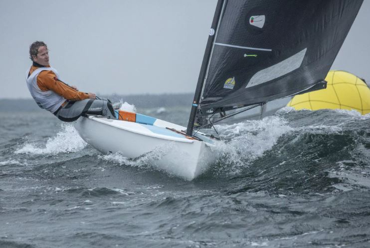 Ein Mann mit einem orangefarbenen Oberteil segelt bei viel Wind eine Jolle mit einem schwarzen Segel daneben ist eine gelbe Tonne.