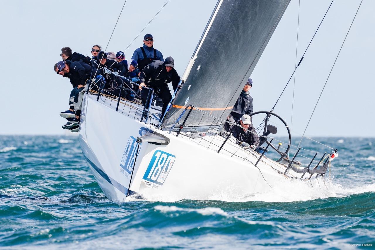 Eine weiße Yacht auf deren Bug die Nummer 18 zu erkennen ist, die Crew trägt schwarzes Ölzeug.