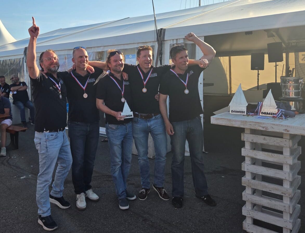 Eine Segelmannschaft mit einem Preis in der Hand reckt jubelnd die Hände in die Höhe, die Männer tragen schwarze Polo-Shirts und haben eine Medaille um den Hals.