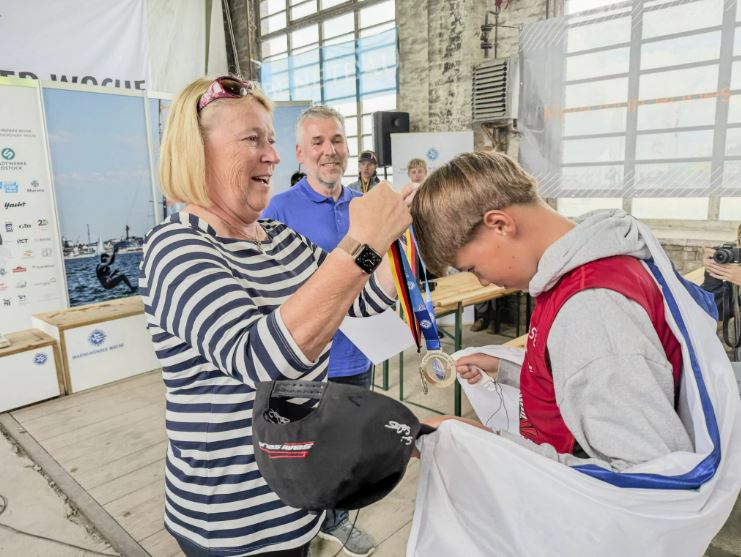 Eine Frau in einem gestreiften T-Shirt hängt einem Jungen eine Medaille für den Sieg bei einem Sportevent um den Hals.