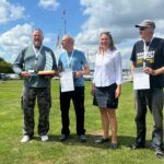 Drei Männer freuen sich bei der Siegerehrung bei einer Segelregatta. Eine Frau gratuliert ihnen.