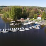 Das Drohnenfoto zeigt einen Steg in einem See, an dem Segelboote liegen. An Land ist ein großes Vereinshaus, dahinter viele Wald.