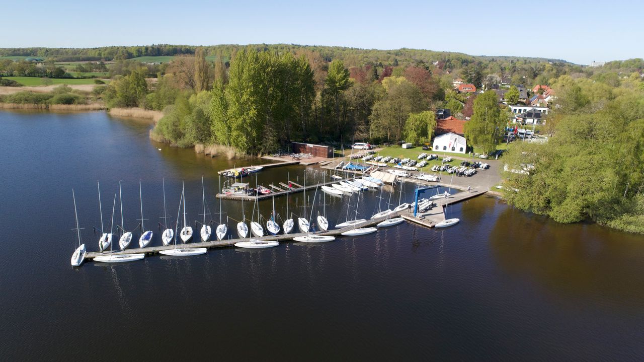 Das Drohnenfoto zeigt einen Steg in einem See, an dem Segelboote liegen. An Land ist ein großes Vereinshaus, dahinter viele Wald.