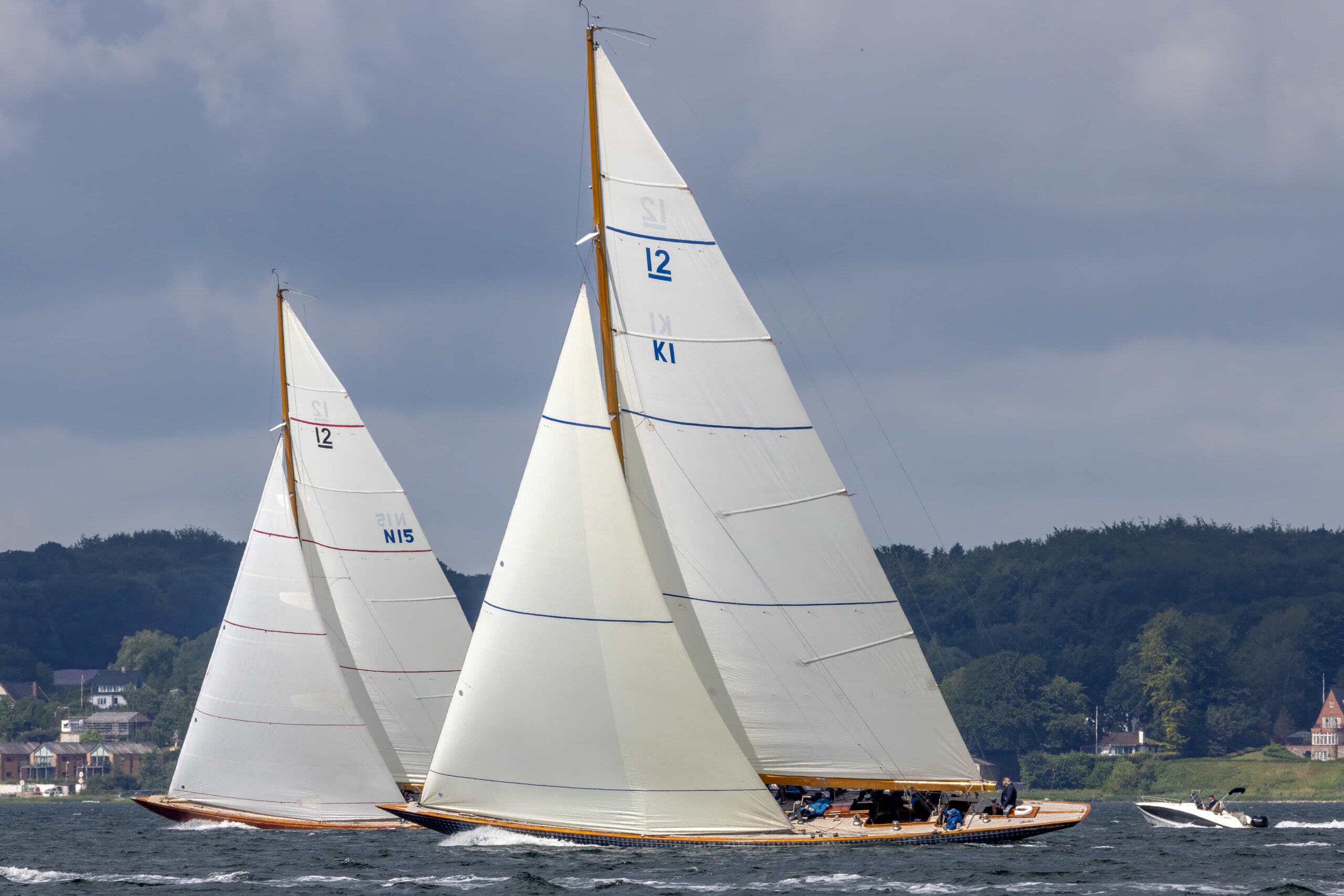 Zwei große klassische Yachten mit einer 12 im Segel vor grauem Himmel und grünen Bäumen.