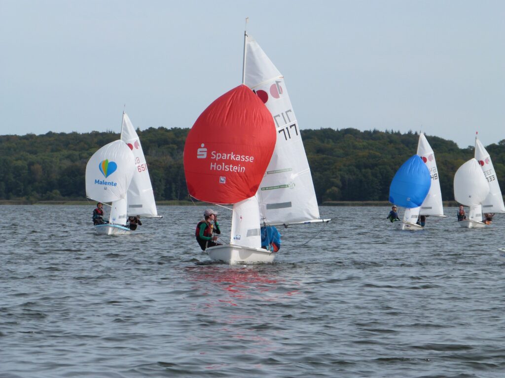 Kleine Boote für Kinder und Jugendliche segeln auf dem Kellersee in Schleswig-Holstein. Vorne ist ein Boot mit einem roten Segel, hinten eines mit einem blauen.