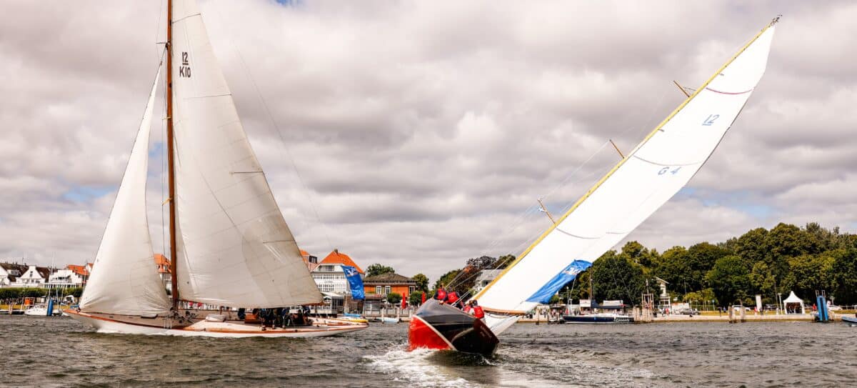 Zwei große Yschten segeln auf der Trave gegeneinander, die eine Yacht hat ein rotes Unterwasserschiff und einen dunkelblauen Rumpf.