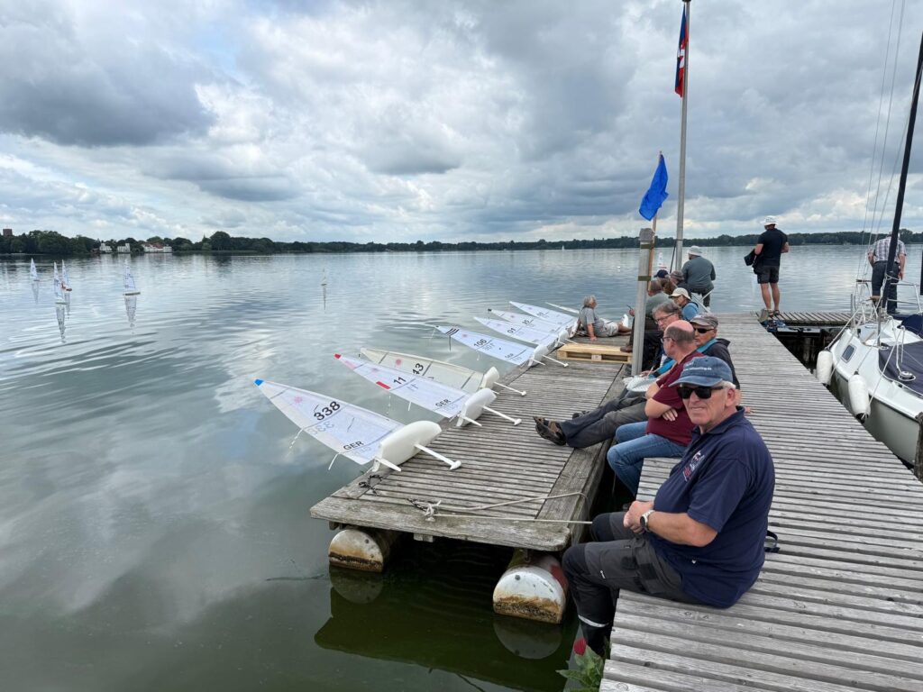 Männer sitzen auf dem Steg. Vor ihnen liegen kleine Modell-Yachten, die ca. einen Meter lang sind. Die Segler warten auf Wind. Das Wasser des Sees ist ruhig.