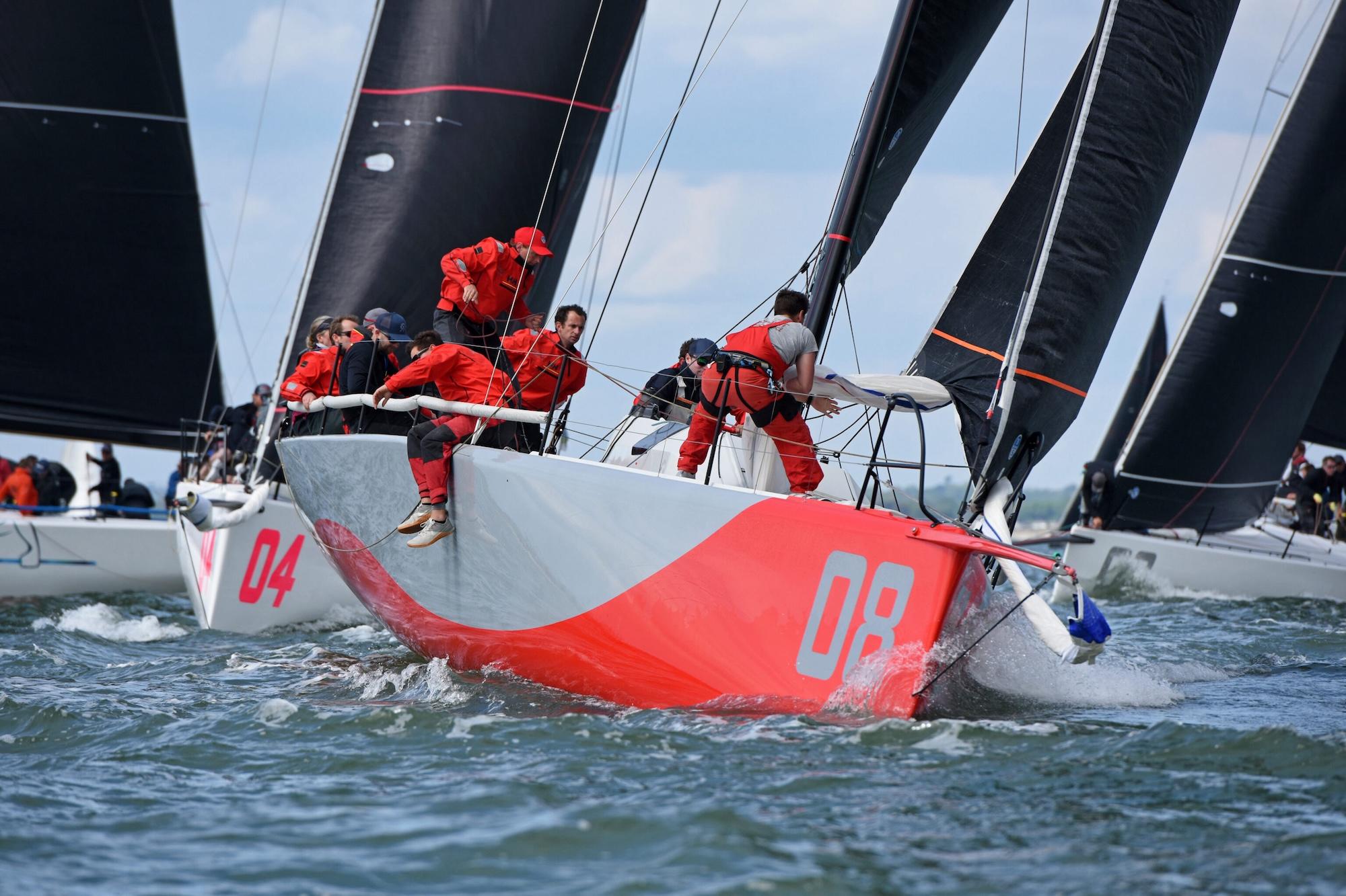 Eine Rennyacht mit einem grau-roten Rumpf und schwarzen Segeln, die Crew trägt rote Bekleidung.