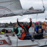 Fröhliche Menschen sitzen auf einer Yacht in der Mitte ein dunkelhäutiger Mann der die Arme hoch hebt