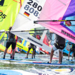 Viel Menschen surfen auf Boards auf einem See. Es gibt gelbe, weiße und pinkfarbene Segel.