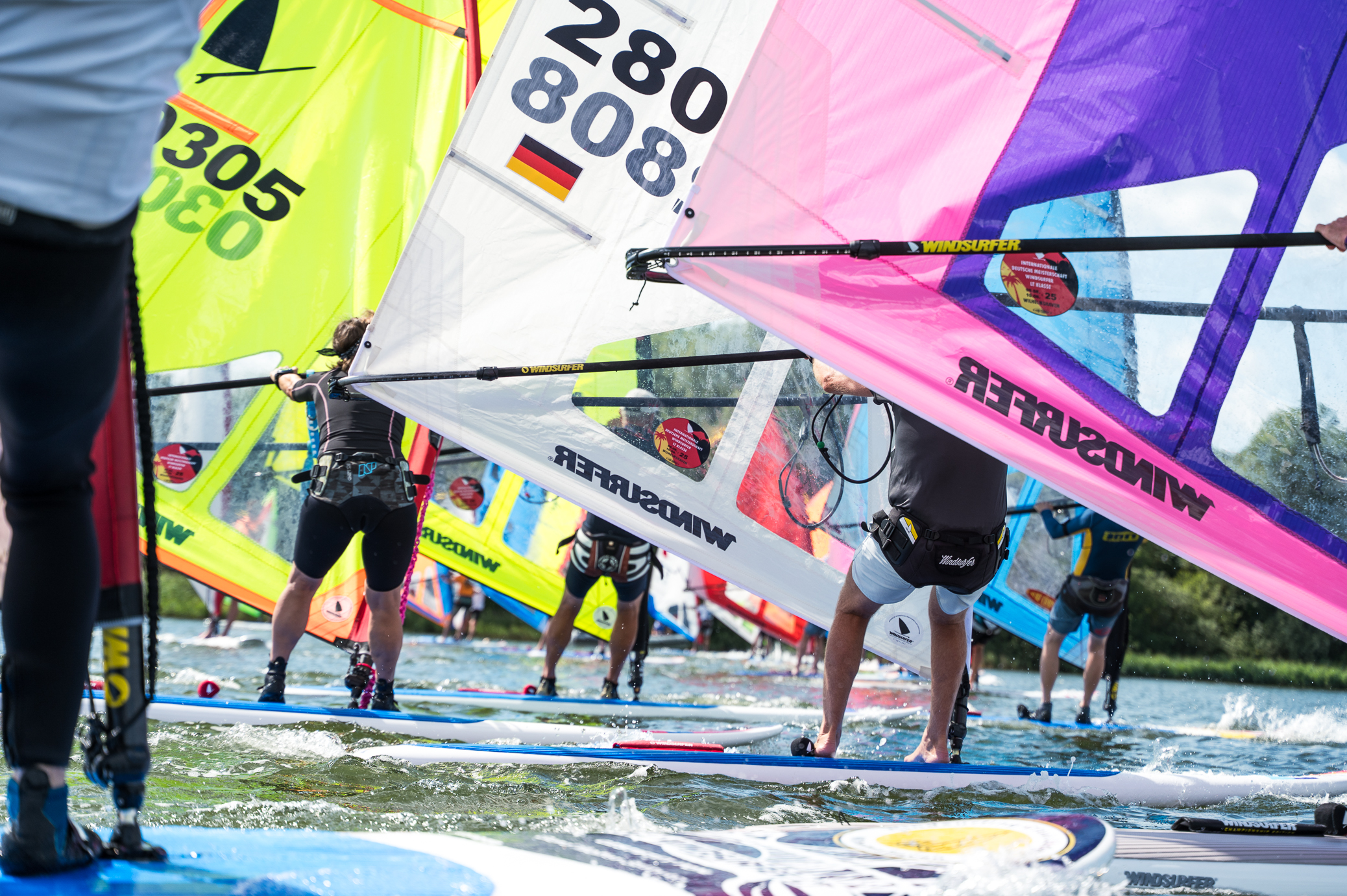 Viel Menschen surfen auf Boards auf einem See. Es gibt gelbe, weiße und pinkfarbene Segel.