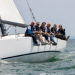 Sechs fröhliche Frauen und Männer segeln auf einer sportlichen Yacht. Das Schiff krängt leicht.