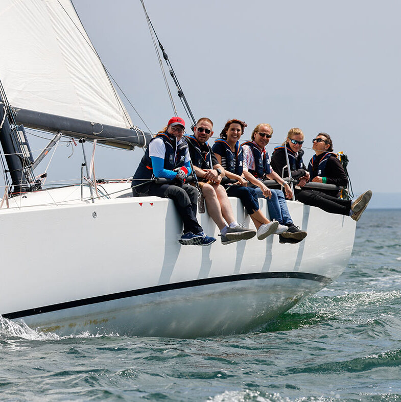 Sechs fröhliche Frauen und Männer segeln auf einer sportlichen Yacht. Das Schiff krängt leicht.