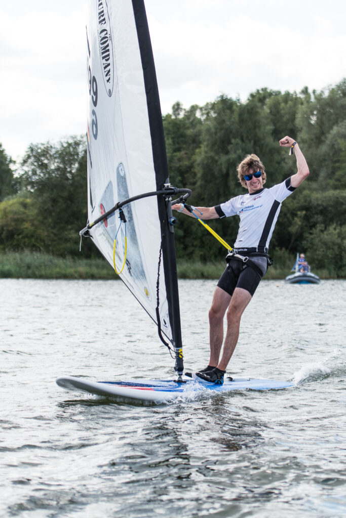 Ein Windsurfer surft auf seinem Board mit einem weißen Segel. Er reckt die Faust in die Höhe und freut sich.