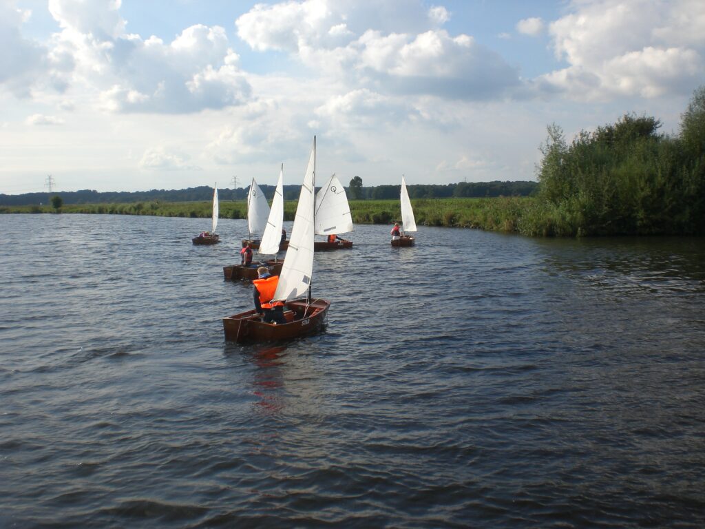 Kinder segeln in kleinen Booten auf einem Fluss. Sie tragen orange-farbene, leuchtende Westen.