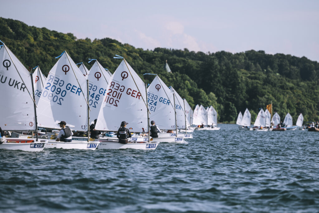 Viele kleine Segelboote warten auf den Start zu einer Wettfahrt. Sie segeln auf einem See. Im Hintergrund ist viel Wald.