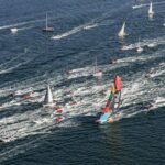 Eine Regattayacht mit bunten Segeln wird von vielen kleinen Motorbooten und einigen Yachten begleitet.