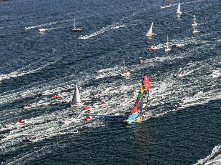 Eine Regattayacht mit bunten Segeln wird von vielen kleinen Motorbooten und einigen Yachten begleitet.