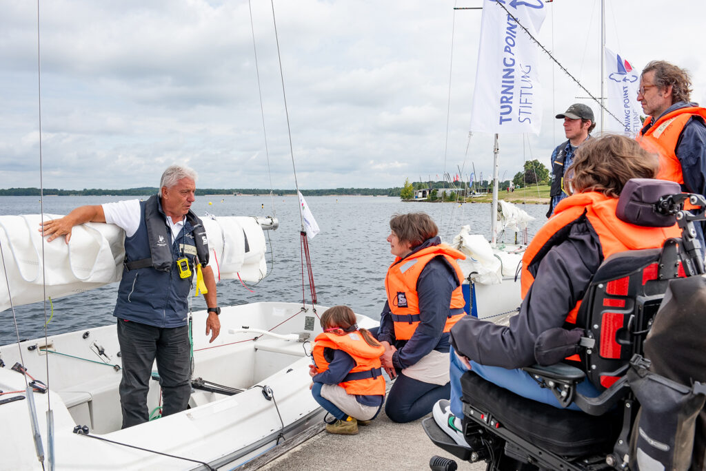 Auf einem Steg stehen Kinder Erwachsen mit Rettungswesten. Sie wollen in das Boot vor ihnen im Wasser steigen.